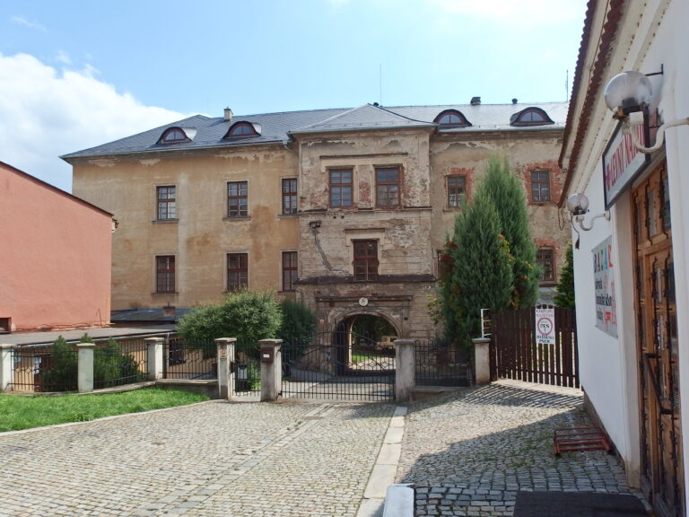 Historická budova v centru Šumperka s oprýskanou fasádou, starobylým arkádovým vstupem a uzavřeným dvorkem, focená z dlažební uličky.